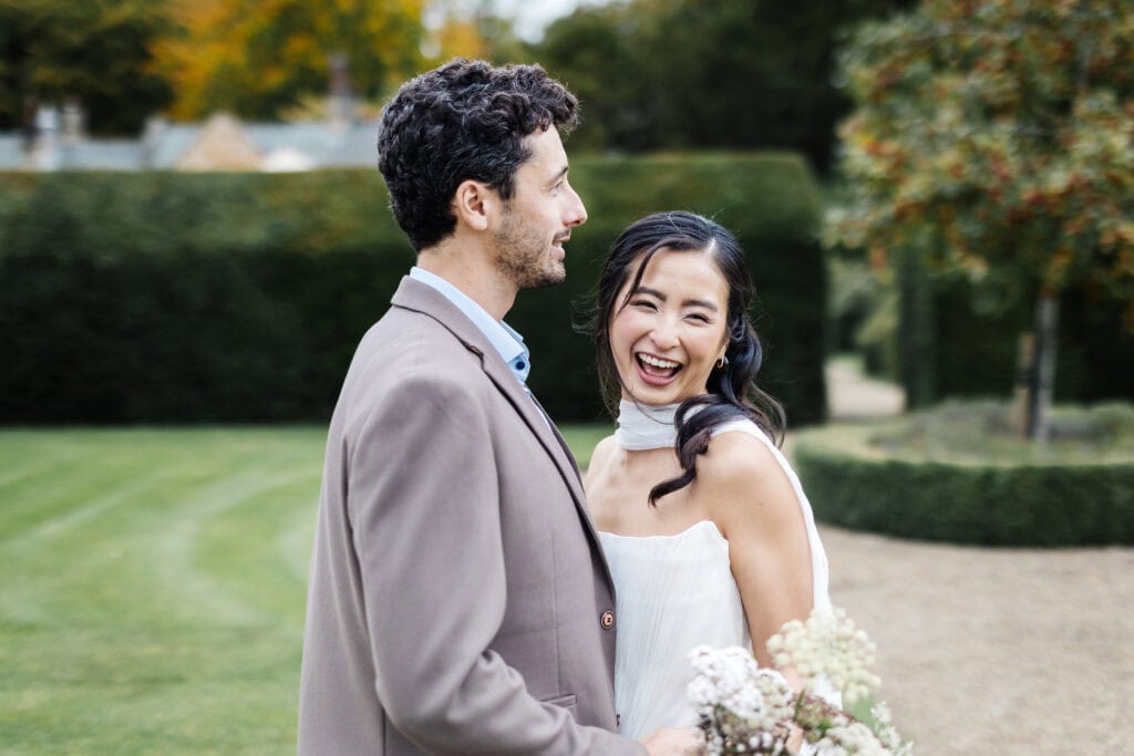 A man in a brown suit and a woman in a white dress stand outdoors, smiling and laughing together, holding a bunch of flowers, with greenery and trees in the background. Drawn by Light Photography