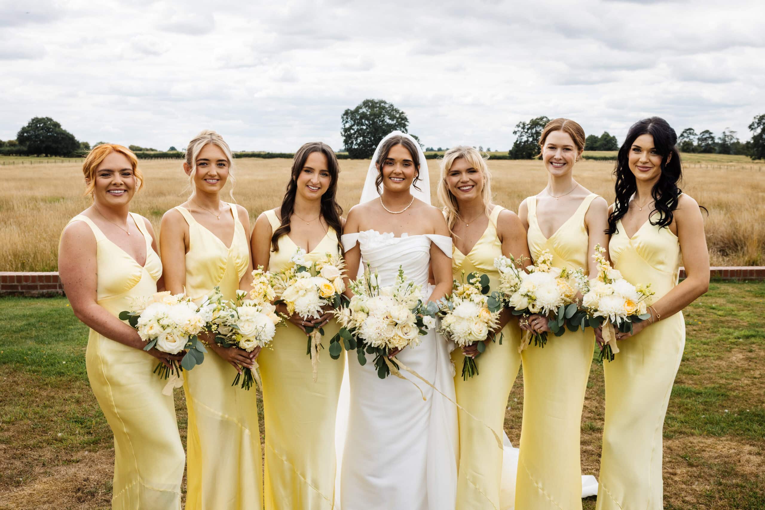 A bride in a white dress stands outdoors at a Middleton Lodge wedding with six bridesmaids in matching yellow dresses, each holding a bouquet of white and yellow flowers. Drawn by Light Photography