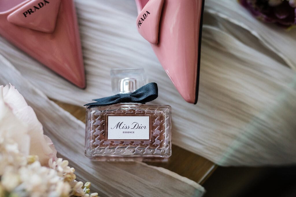 A bottle of Miss Dior Essence perfume with a black bow is placed near a pair of pink Prada shoes on a light fabric background, evoking the elegance of a Middleton Lodge wedding. Drawn by Light Photography