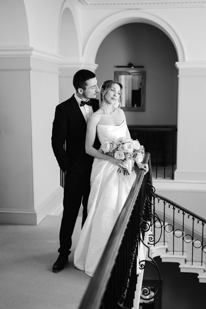 A bride and groom stand together on an elegant indoor balcony at their Middleton Lodge wedding. The bride holds a bouquet whilst the groom stands behind her, both facing slightly to the side in a beautifully formal setting. Drawn by Light Photography