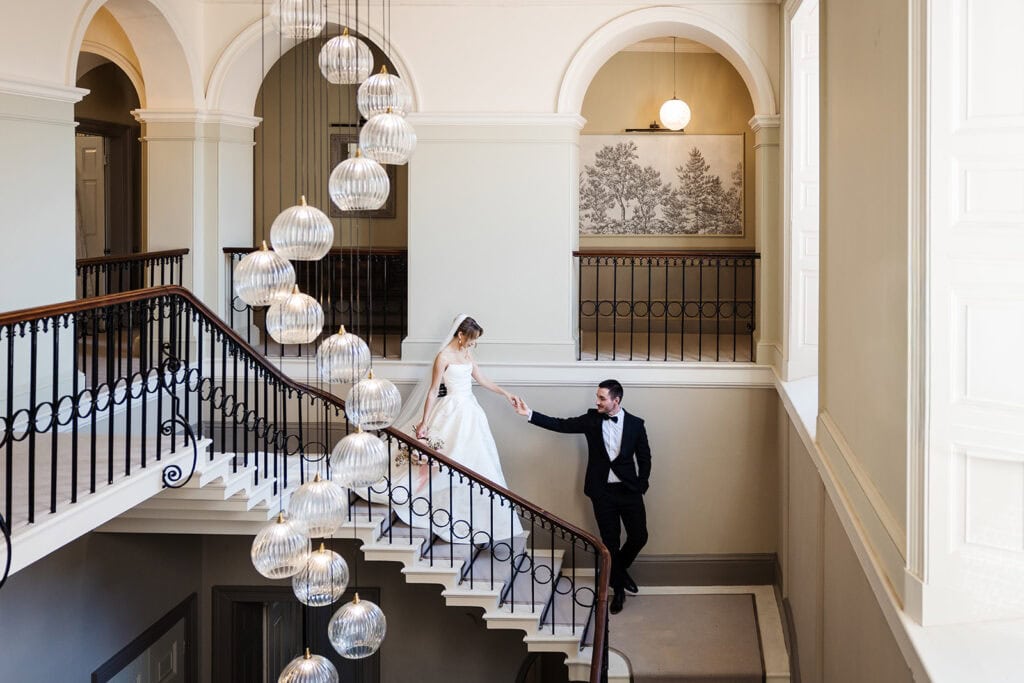 A bride and groom stand on a grand staircase inside the bright, elegant Middleton Lodge wedding venue, with large windows and spherical hanging lights. Drawn by Light Photography
