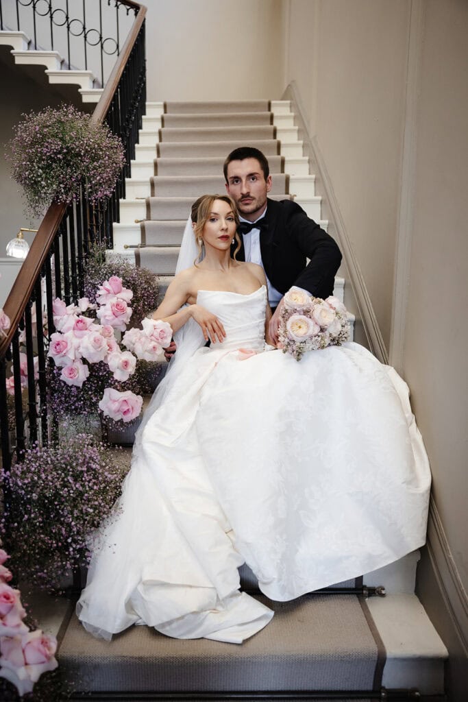 A bride and groom sit together on a staircase decorated with pink flowers at their Middleton Lodge wedding, the bride in a white dress and the groom in a black dinner suit, both looking at the camera. Drawn by Light Photography