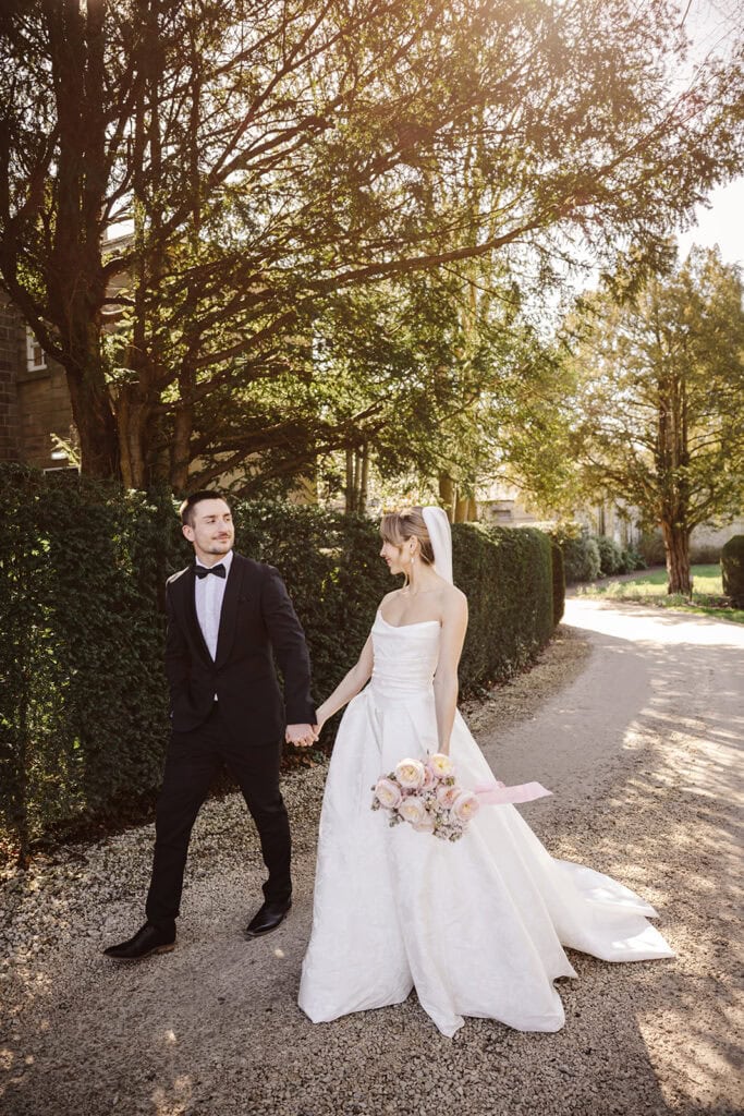 A bride in a white gown and a groom in a black suit walk hand in hand on a gravel path at their Middleton Lodge wedding, surrounded by greenery and sunlight. Drawn by Light Photography