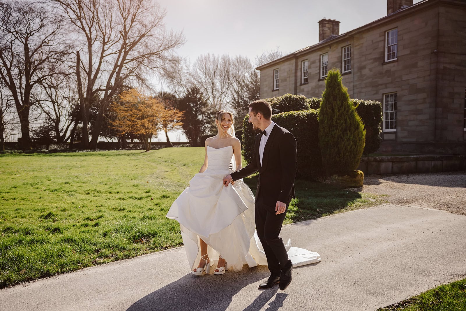 A bride and groom walk together outdoors on a sunny day near the historic stone building at Middleton Lodge, with the bride holding her dress. Drawn by Light Photography