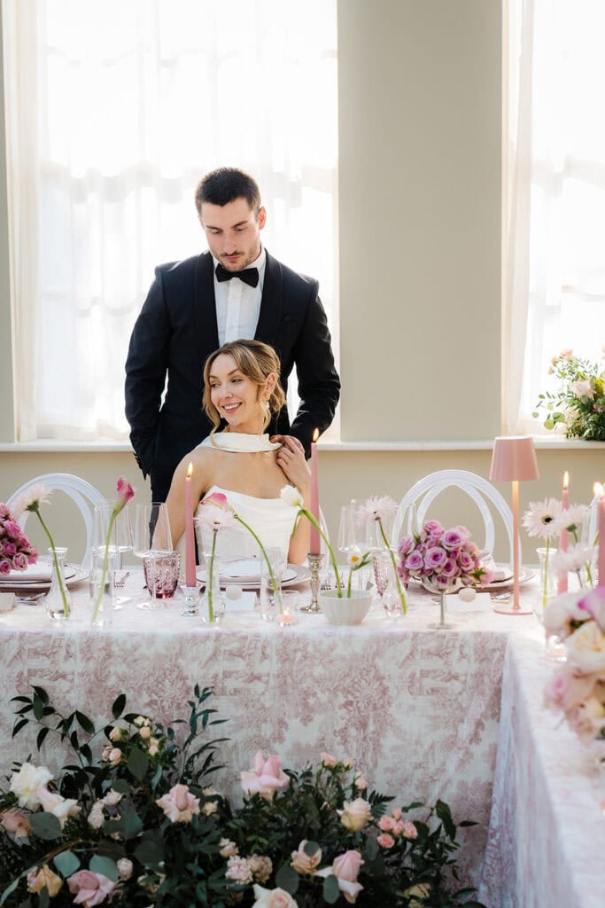 A man in a black suit stands behind a seated woman in a white dress at a beautifully decorated Middleton Lodge wedding table with pink and white flowers and candles. Drawn by Light Photography
