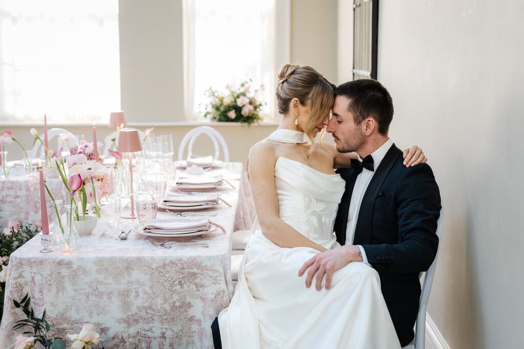 A bride in a white dress sits on the lap of a groom in a black suit at a beautifully decorated Middleton Lodge wedding table, sharing an intimate moment. Drawn by Light Photography
