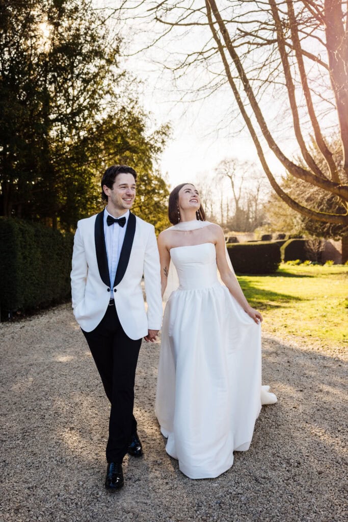 A bride in a white strapless gown and a groom in a white tuxedo jacket walk hand in hand outdoors on a gravel path, surrounded by greenery and sunlight. Drawn by Light Photography