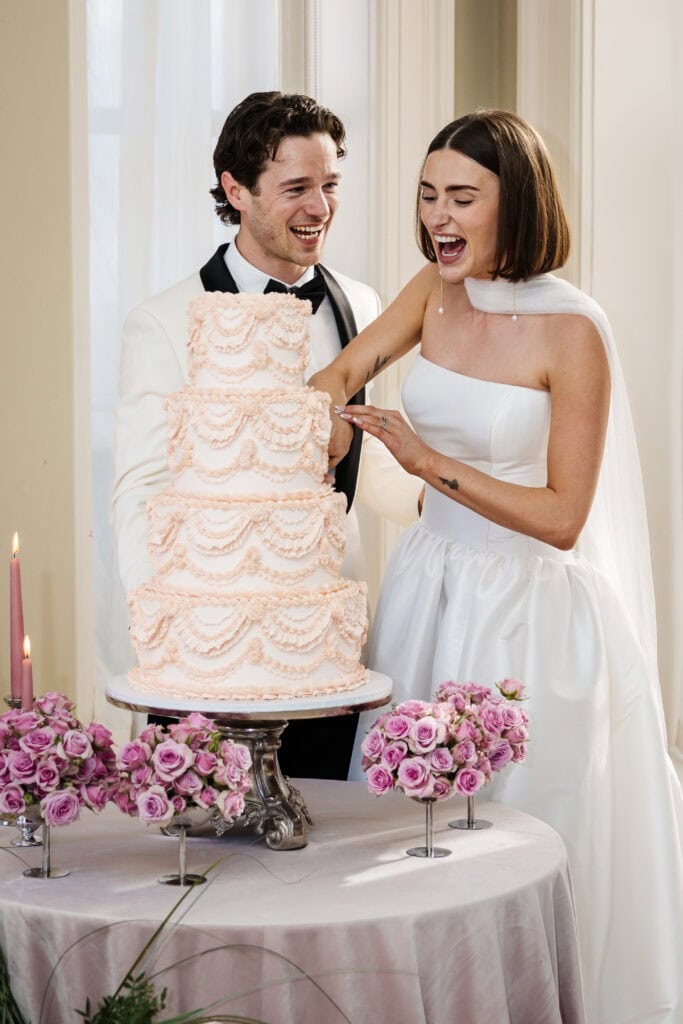 A bride and groom, both in formal attire, smile and laugh together as they cut a tall, ornate wedding cake at a decorated table with pink flowers and candles. Drawn by Light Photography