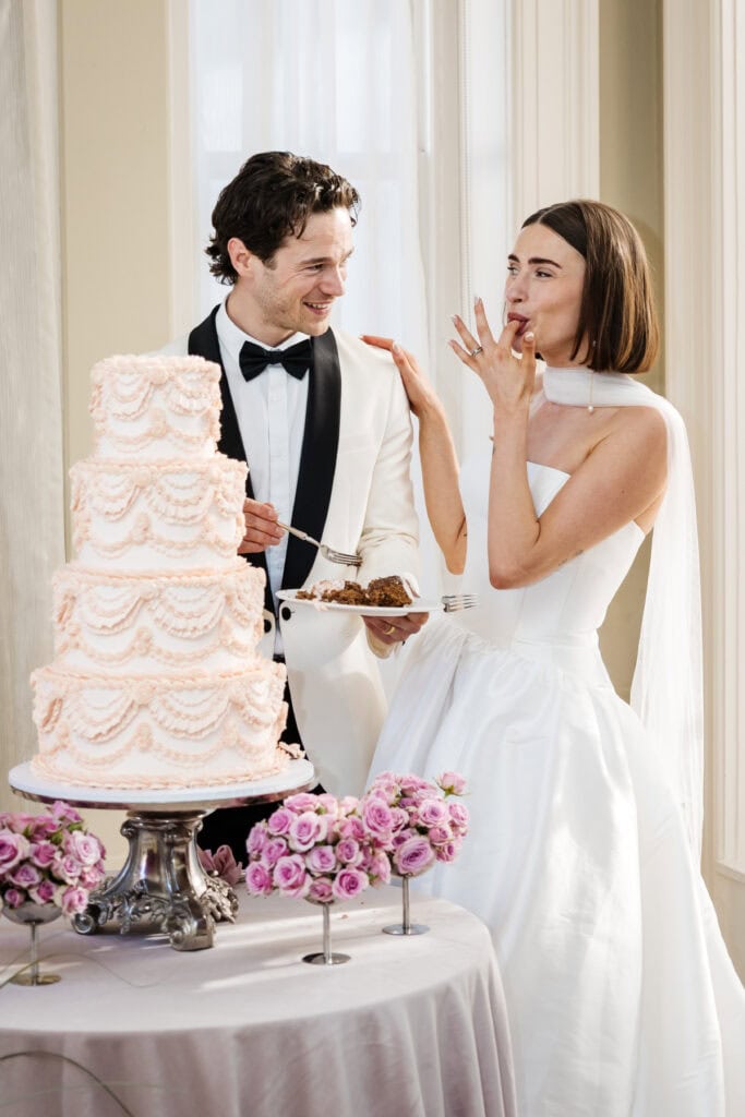A bride and groom in formal attire stand by a tiered wedding cake; the bride tastes icing from her finger while the groom holds a plate with a slice of cake. Drawn by Light Photography