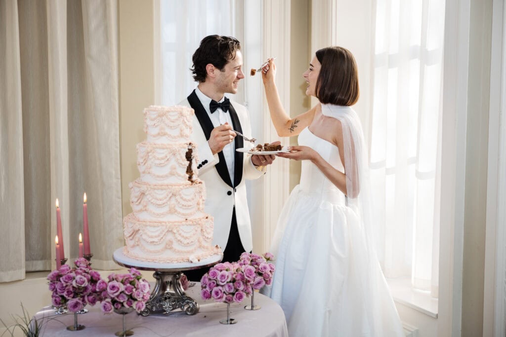 A bride and groom in formal attire feed each other cake beside a decorated tiered wedding cake on a table with pink flowers and candles. Drawn by Light Photography