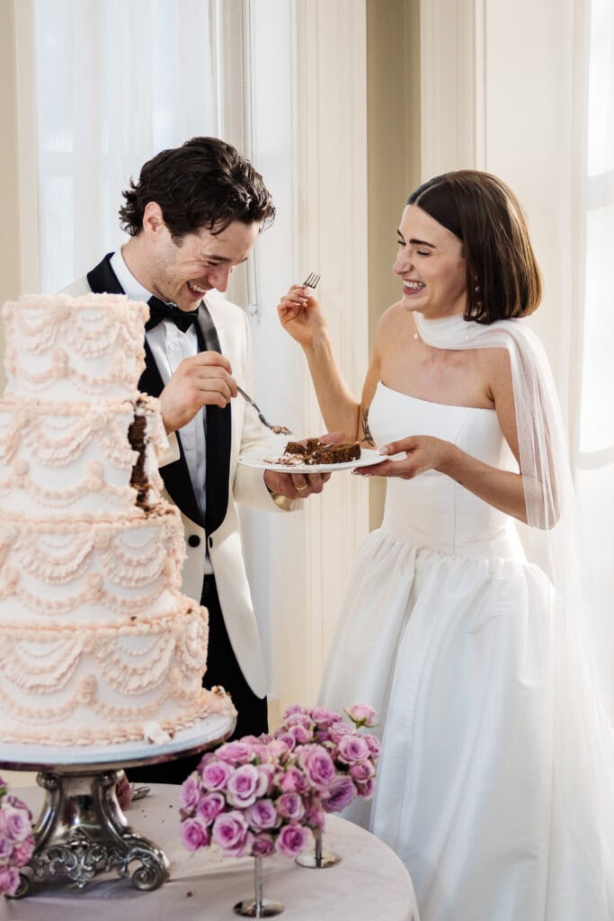 A bride and groom in formal attire smile and eat cake together next to a decorated tiered wedding cake and a bouquet of purple roses. Drawn by Light Photography