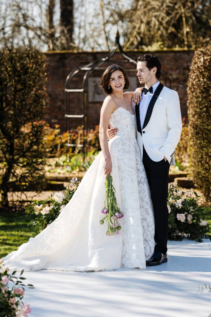 Bride in a white gown holding flowers stands next to groom in a white tuxedo jacket; both pose outdoors on a sunny day surrounded by greenery and floral arrangements. Drawn by Light Photography
