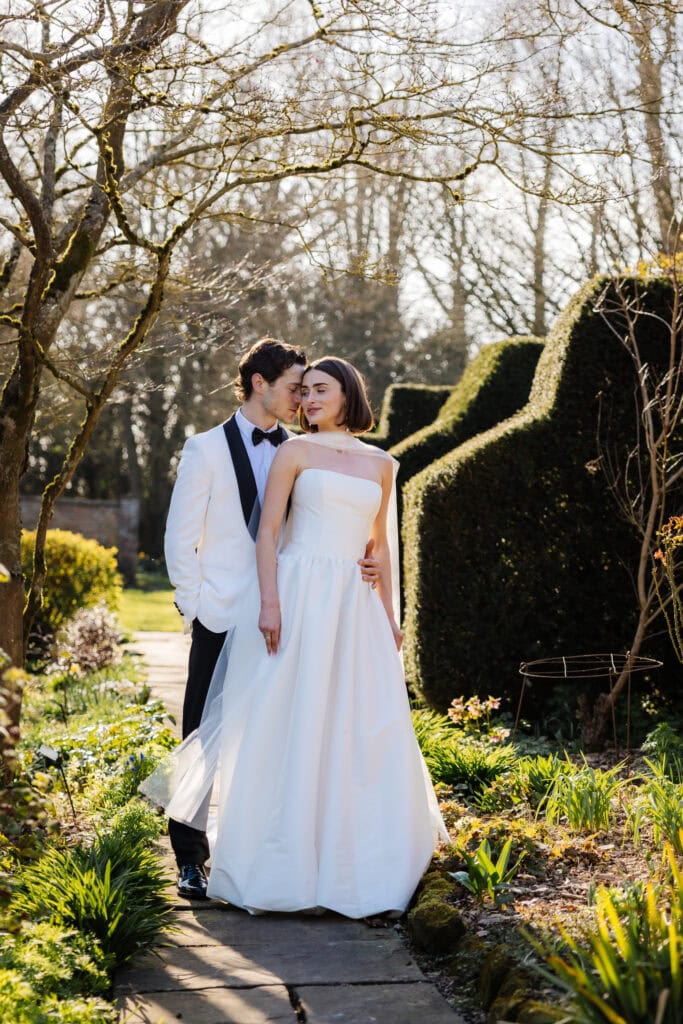 A bride and groom stand together on a garden path, with the groom in a white dinner jacket and the bride in a strapless white dress, surrounded by greenery and sunlight. Drawn by Light Photography