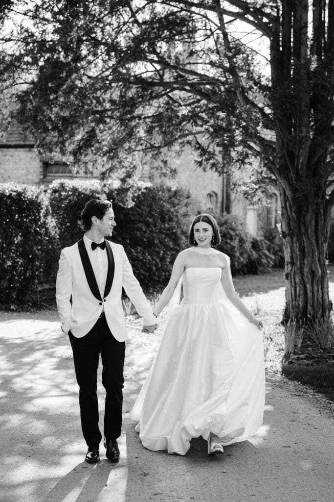 A couple in formal wedding attire walk hand in hand outdoors, with trees and a building in the background. Drawn by Light Photography