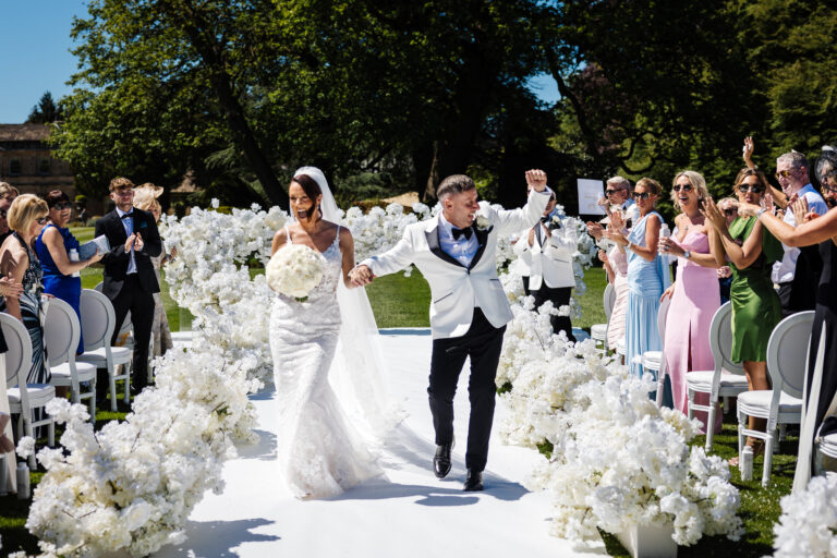 A bride and groom walk joyfully down an outdoor aisle lined with white flowers at their Middleton Lodge wedding as guests applaud on a sunny day. Drawn by Light Photography