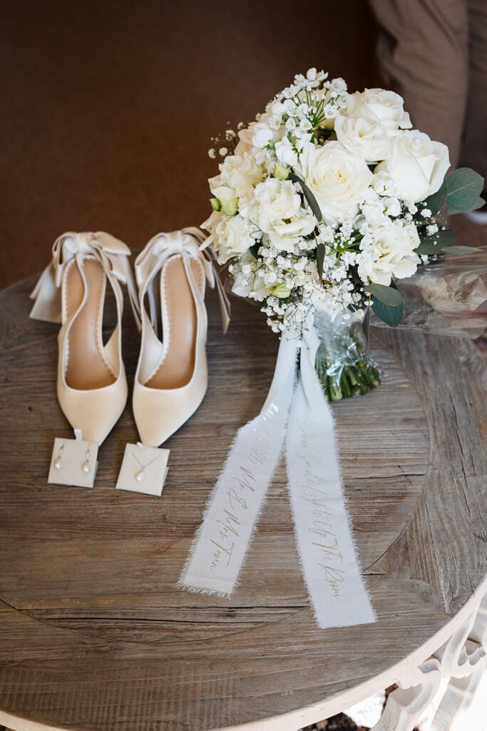 A pair of white bridal shoes, pearl earrings, and a bouquet of white flowers with greenery arranged on a round wooden table. Drawn by Light Photography