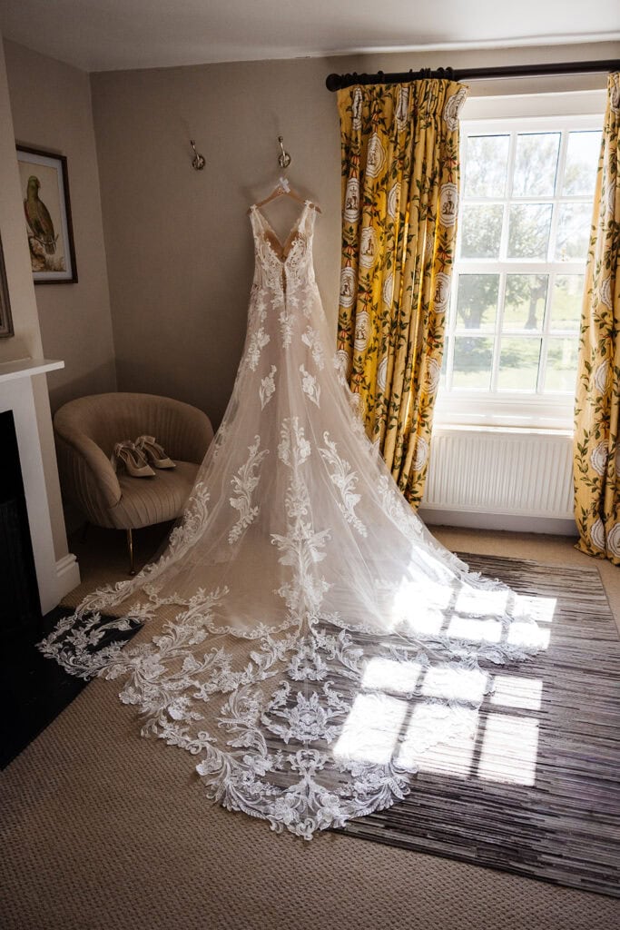 A lace wedding dress with a long, detailed train hangs on a wall in a sunlit room next to a window with yellow curtains. Drawn by Light Photography