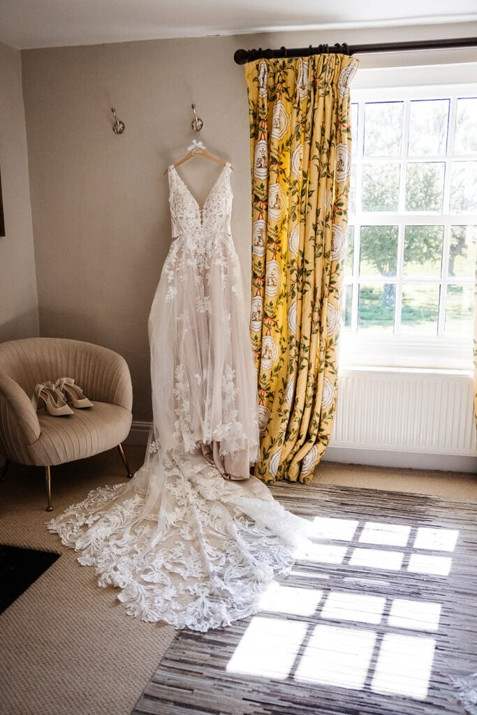 A white lace wedding dress with a long train hangs on a wall next to a window with yellow patterned curtains and an armchair with a small bag. Drawn by Light Photography