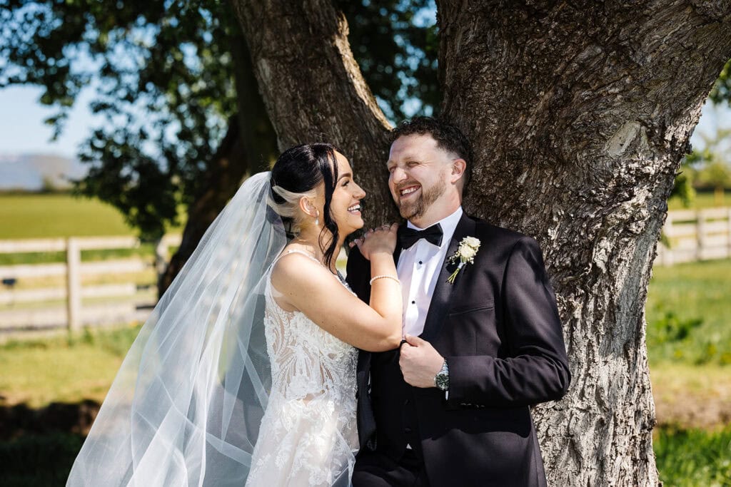 A bride and groom stand together smiling and laughing under a tree outdoors, with the bride touching the groom’s lapel. Drawn by Light Photography