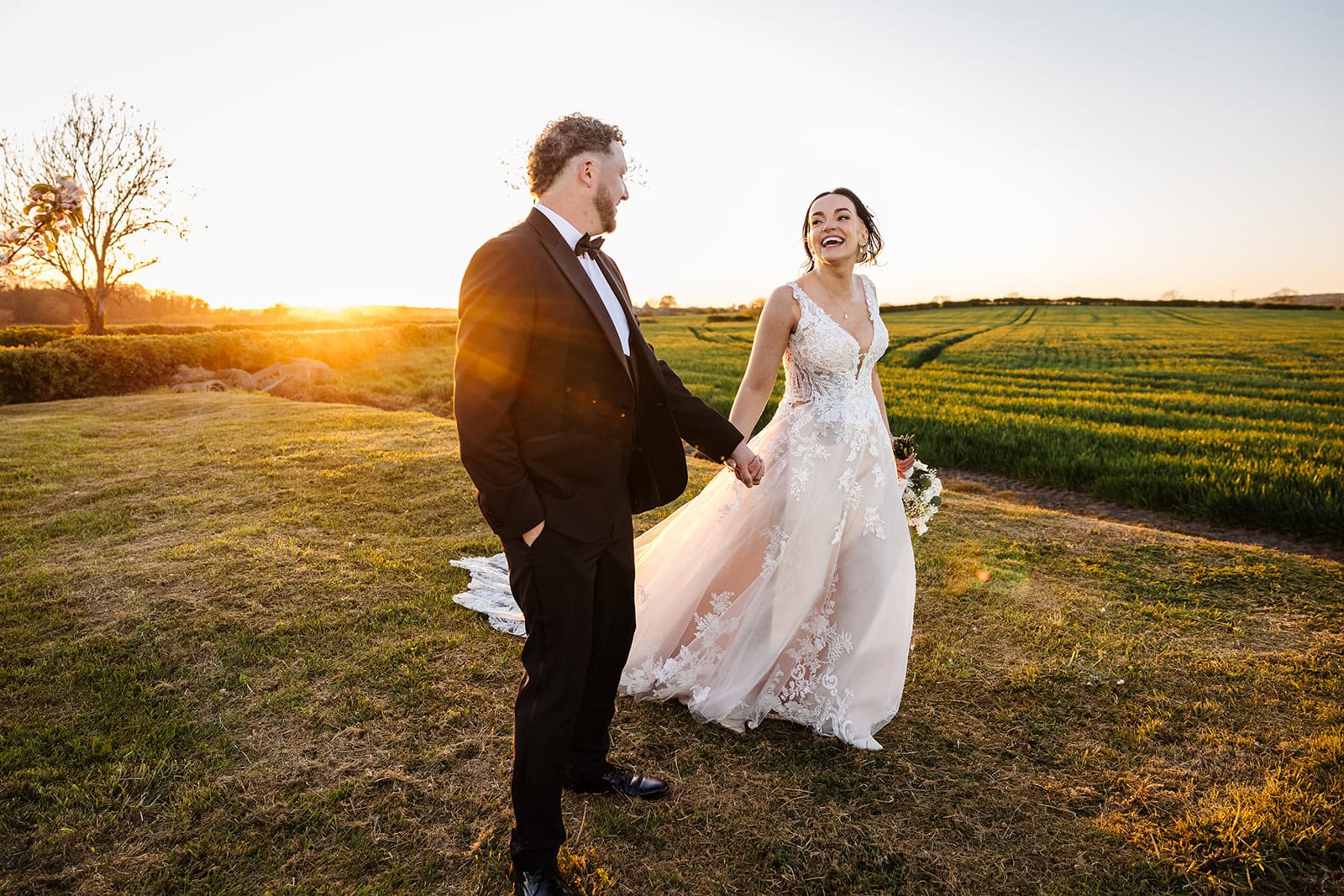 A bride and groom hold hands and walk outdoors in a grassy field at sunset, both smiling and dressed in formal wedding attire. Drawn by Light Photography
